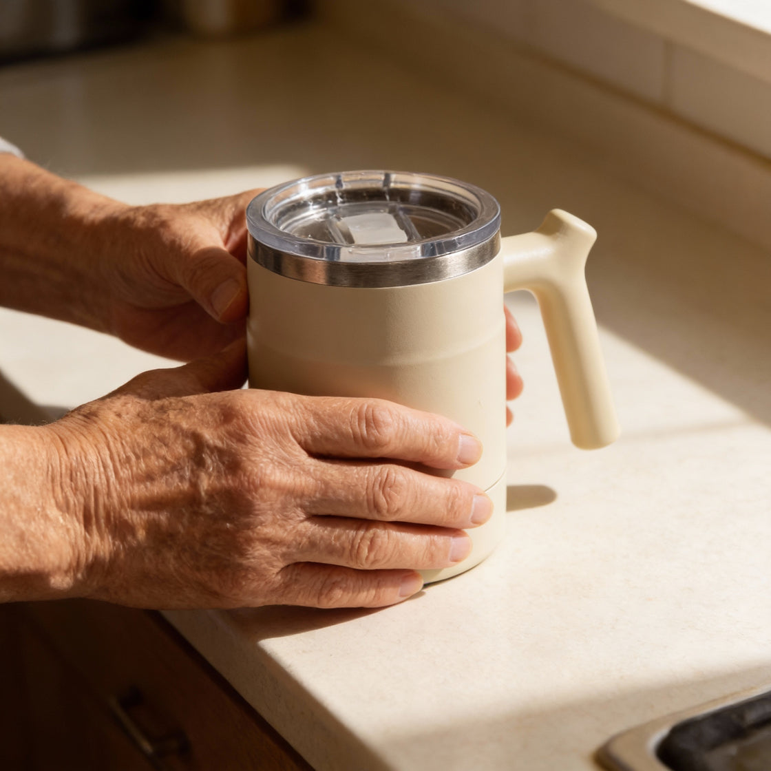 Older hands holding an insulated easy-grip cup on a kitchen bench